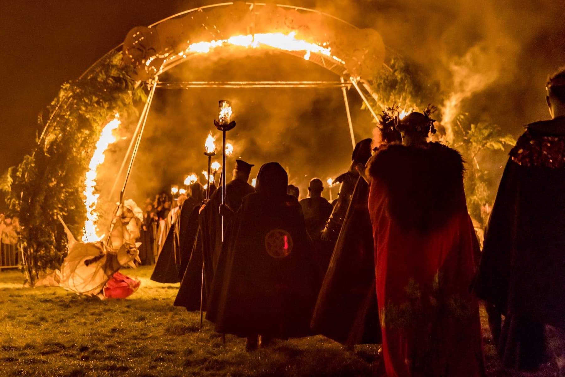 Group of people wearing various hooded costumes marching through and archway of fire carrying medieval torches
