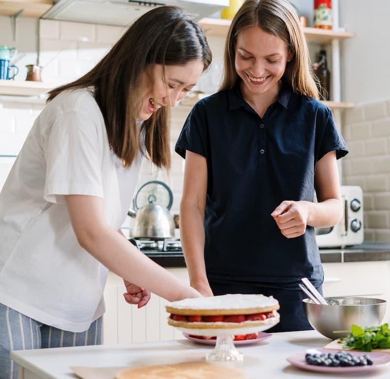 2 women laughing as they assemble layers of sponge cake and berries
